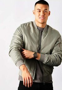 Khakifarbenes Bomberjacke mit gerippten Bündchen und Kragen, mit einem Reißverschlussfach am Arm, getragen über einem grauen T-Shirt. Modell trägt eine dunkle Armbanduhr.