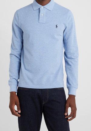 Homme portant un polo à manches longues bleu clair avec deux boutons blancs et un jean bleu foncé, debout devant un fond uni.