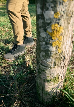 Černé kotníkové boty z texturované semišové kůže s gumovou podrážkou, spárované s khaki kalhotami, stojící blízko kmeni stromu pokrytého lišejníky.