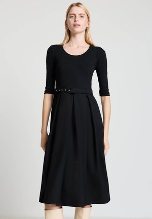 Femme portant une robe noire ceinturée à manches trois-quarts et des bottes beige montant jusqu'aux genoux, debout devant un fond clair uni.