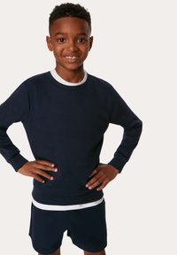 Sudadera azul marino con puños y dobladillo acanalados, llevada sobre una camiseta blanca de mangas cortas. La modelo está de pie con las manos en las caderas, sonriendo.