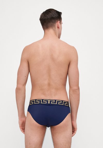 Marine svømmebriefs med et gullig geometrisk mønster på midjen; glatt stoff; tettsittende design som former seg til kroppen.