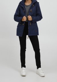 Marinblå vattentät jacka med dragkedja och dragsko, med två framfickor och avtagbar huva. Bärs över en grå tröja och svarta byxor.