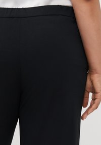 Close-up of a person’s waist and hand wearing black elastic waistband pants and a white top against a plain background.