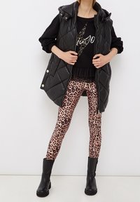 Chaleco acolchado negro con capucha, usado sobre una blusa negra con dobladillo con volantes, combinado con leggins de estampado de leopardo y botines negros.