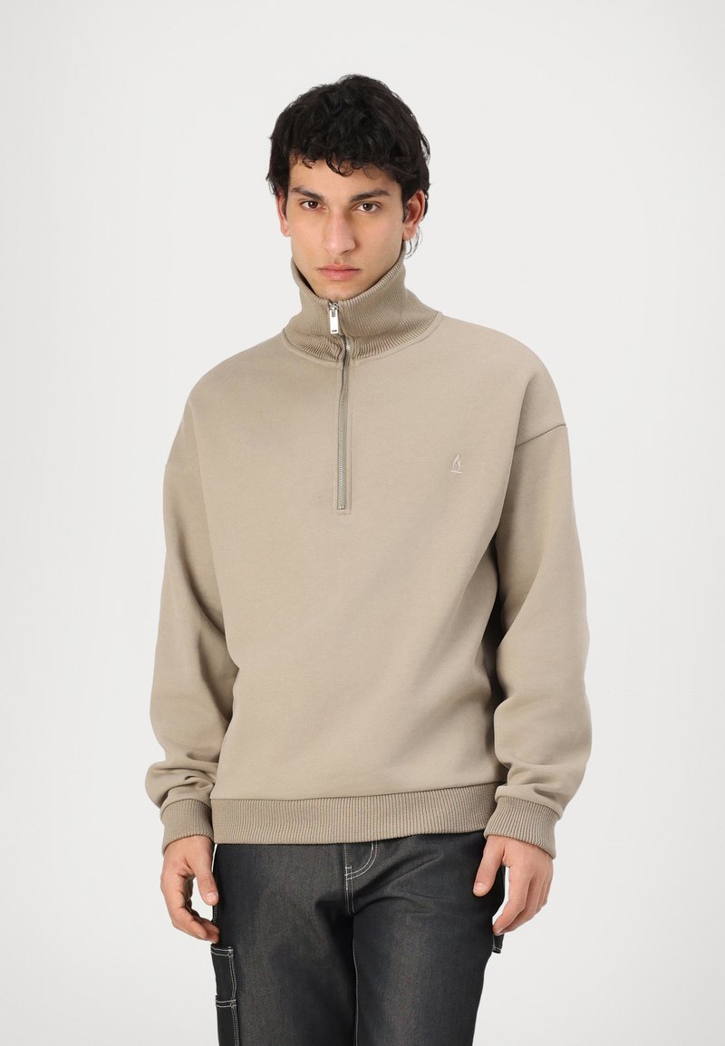 Sudadera beige con cremallera y cuello alto, puños y dobladillo acanalados. Presenta un pequeño logo en el pecho. Se lleva con jeans oscuros.