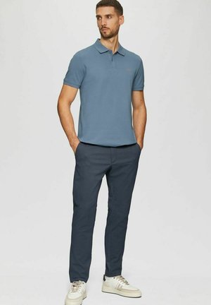 Mann steht mit den Händen in den Taschen, trägt ein blaues kurzärmeliges Poloshirt, dunkle Hose und weiße Turnschuhe vor einem einfarbigen Hintergrund.