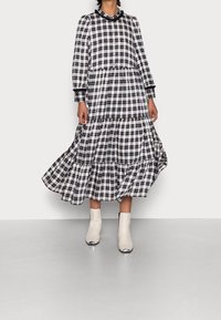 Černobíle kostkovaná dlouhá šaty s vrstvenými volány, dlouhými rukávy, knoflíkovým zapínáním a černými detaily; doplněné bílými kotníkovými botami.