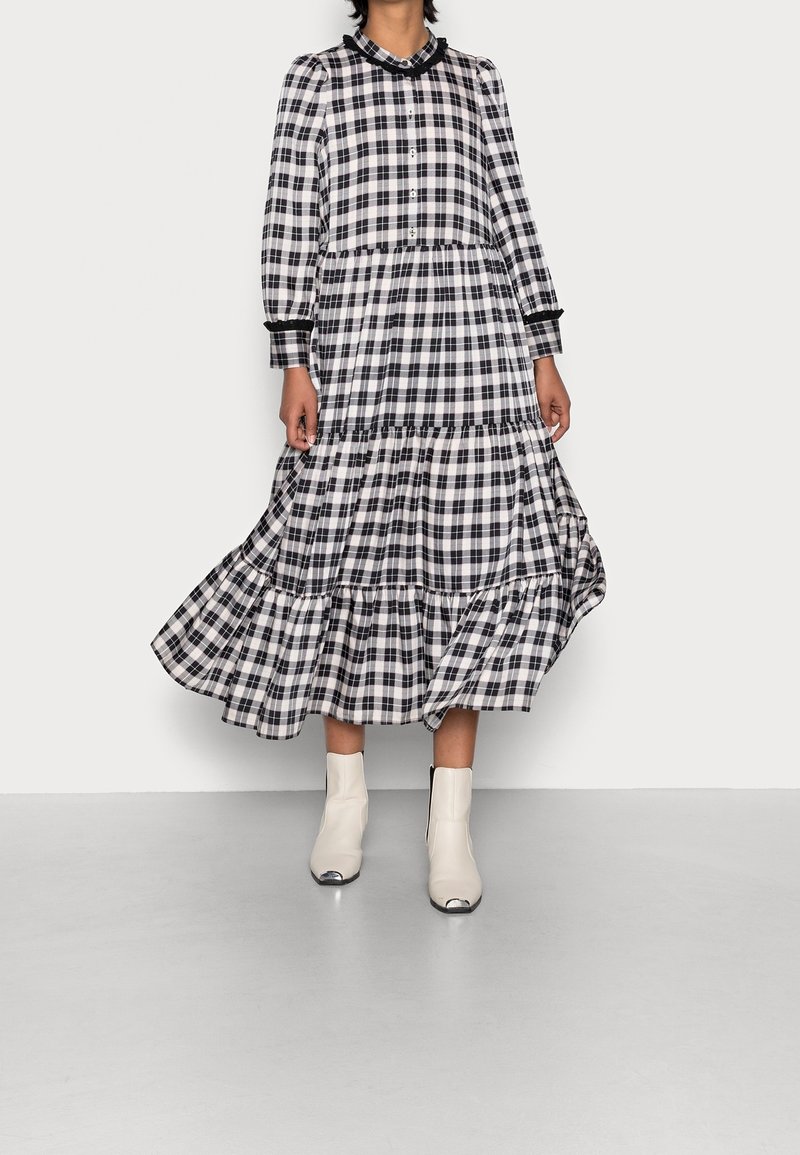 Černobíle kostkovaná dlouhá šaty s vrstvenými volány, dlouhými rukávy, knoflíkovým zapínáním a černými detaily; doplněné bílými kotníkovými botami.
