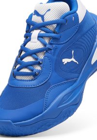 Zapatilla deportiva azul con materiales de malla y cuero sintético, que presenta una superficie texturizada, acentos en blanco y un logotipo prominente. Diseño con cordones.