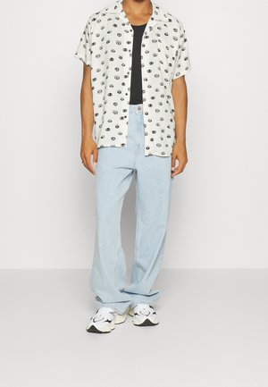 Camicia a maniche corte con bottoni, motivo di occhi neri su sfondo chiaro, abbinata a jeans a gamba larga azzurri e sneakers con motivo.