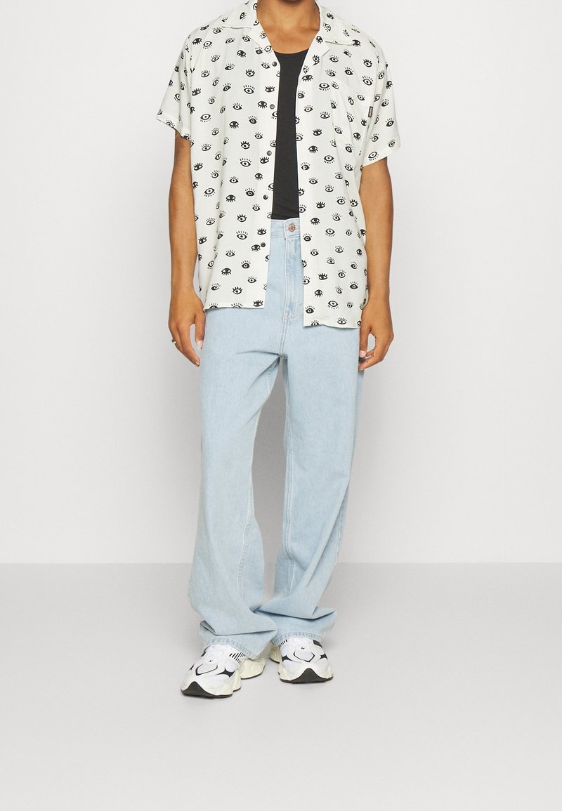 Kurzärmeliges Hemd mit schwarzen Augen-Muster auf hellem Hintergrund, kombiniert mit hellblauen Jeans mit weitem Bein und gemusterten Sneakers.