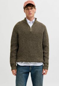Khaki-grüner Strickpullover mit einem halben Reißverschlusskragen, gerippter Textur, langen Ärmeln, getragen über einem weißen Hemd mit Kragen und blauen Jeans.