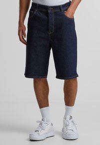 Shorts en denim bleu foncé avec une coupe droite, dotés de poches et de coutures jaunes. Associés à des baskets blanches et des chaussettes.