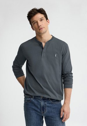 Młody mężczyzna o ciemnych włosach, ubrany w ciemnoszarą długorękawową koszulkę henley i niebieskie dżinsy, stoi na tle jednolitego jasnego tła.