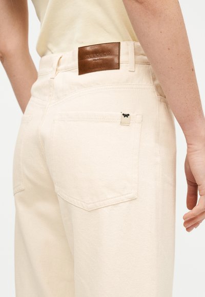 Πλάγια όψη δεξιάς πλευράς ενός ατόμου που φοράει τζιν εκρού με ένα καφέ δερμάτινο ετικέτα που γράφει "WEEKEND MaxMara" πάνω από την πίσω τσέπη.