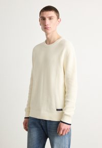 Sweter w kolorze kremowym, dziany, z okrągłym dekoltem, długimi rękawami i żebrową fakturą; posiada metkę w dolnym lewym rogu.