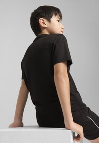 T-shirt en coton noir à manches courtes, avec un col rond. L'enfant est assis sur un piédestal blanc, tourné légèrement sur le côté.