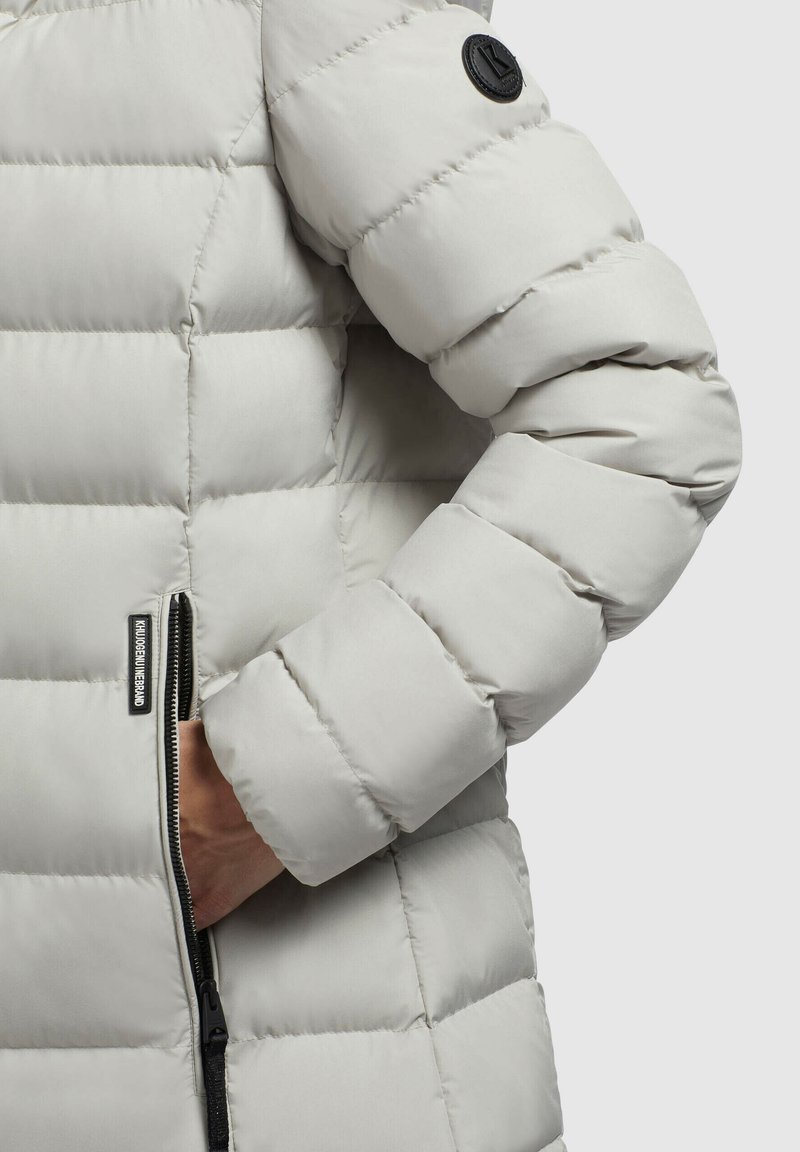 Hellegraue Pufferjacke mit großen gesteppten Abschnitten, einem seitlichen Reißverschlusstaschen und einem runden schwarzen Logo-Patch am Ärmelsaum. Glatte Textur.