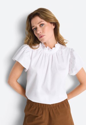 Kobieta o jasnobrązowych włosach, ubrana w białą bluzkę z falbanami i brązowe spodnie z elastycznym pasem, stojąca na tle białego tła.