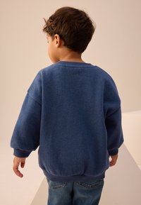 Kék pulóver kerek nyakkivágással és laza, puffadt ujjakkal. Az anyaga puhanak és texturáltnak tűnik, farmernadrággal párosítva.