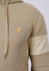 Beige hoodie van zachte katoen met een kangoeroezak, voorzien van een gouden geborduurd vogellogo op de borst en lichtbeige kleurblokken accents.