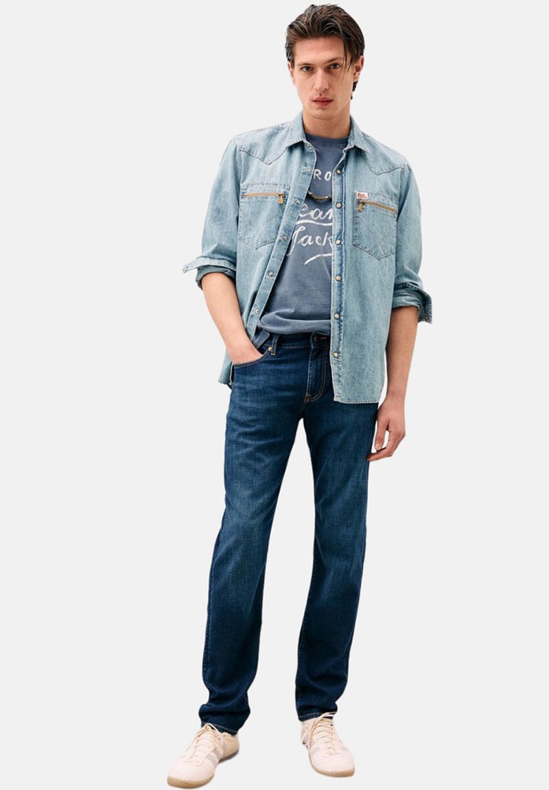 Camicia in denim azzurro chiaro con due tasche sul petto, t-shirt grigia con grafica sotto, jeans blu scuro e sneakers chiare.