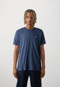 Camiseta de algodón azul marino con cuello redondo y mangas cortas, que presenta un pequeño logo en el pecho. Textura suave y ajuste relajado.