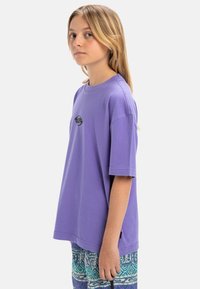 Camiseta de algodón púrpura con mangas cortas, escote redondo y pequeño logotipo en el pecho. La tela tiene una textura suave.