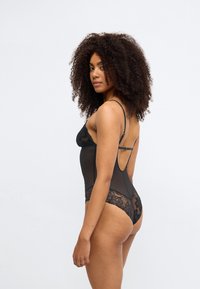 Melnas bodijs ar mežģīņu akcentiem, caurspīdīgu tīkliņa auduma daļu, regulējamām lencēm un zemu aizmugures dizainu, izceļot smalkas detaļas.