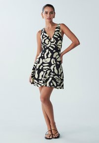 Sort og cremefarvet mønstret kjole med dyb V-udskæring, ærmeløs design, udvidet skørt og letvægtsstof. Bærres med enkle sorte sandaler.