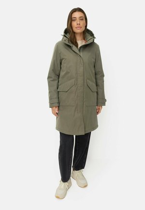 Kvinde iført knælængde olivengrøn hættetrøje, sorte jeans og beige sneakers står foran en ensfarvet lys baggrund.