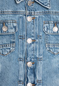 Nahaufnahme einer hellblauen Denimjacke vorne mit Metallknöpfen und zwei aufgesetzten Brusttaschen mit orangenen Nähten.