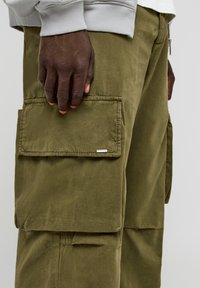 Pantalon cargo en coton vert olive avec de grandes poches latérales, des coutures visibles et une petite étiquette de marque sur la poche. Tissu texturé.