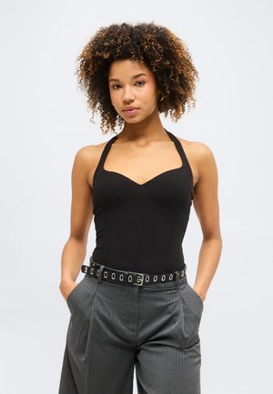 Mladá žena s kučeravými vlasmi, ktorá má na sebe čierny halter-top, sivé nohavice s tenkými pruhmi a čierny opasok so striebornými očkami, ruky v kapsách.