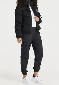 Svart uppblåsbar jacka med hög krage, dragkedjestängning och elastiska ärmslut, i kombination med svarta cargo-byxor med flera fickor och snörning vid anklarna.