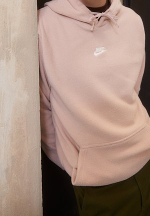 Osoba nosící světle růžovou mikinu Nike s přední kapsou, stojící vedle strukturované béžové stěny, dolní část obličeje není viditelná.