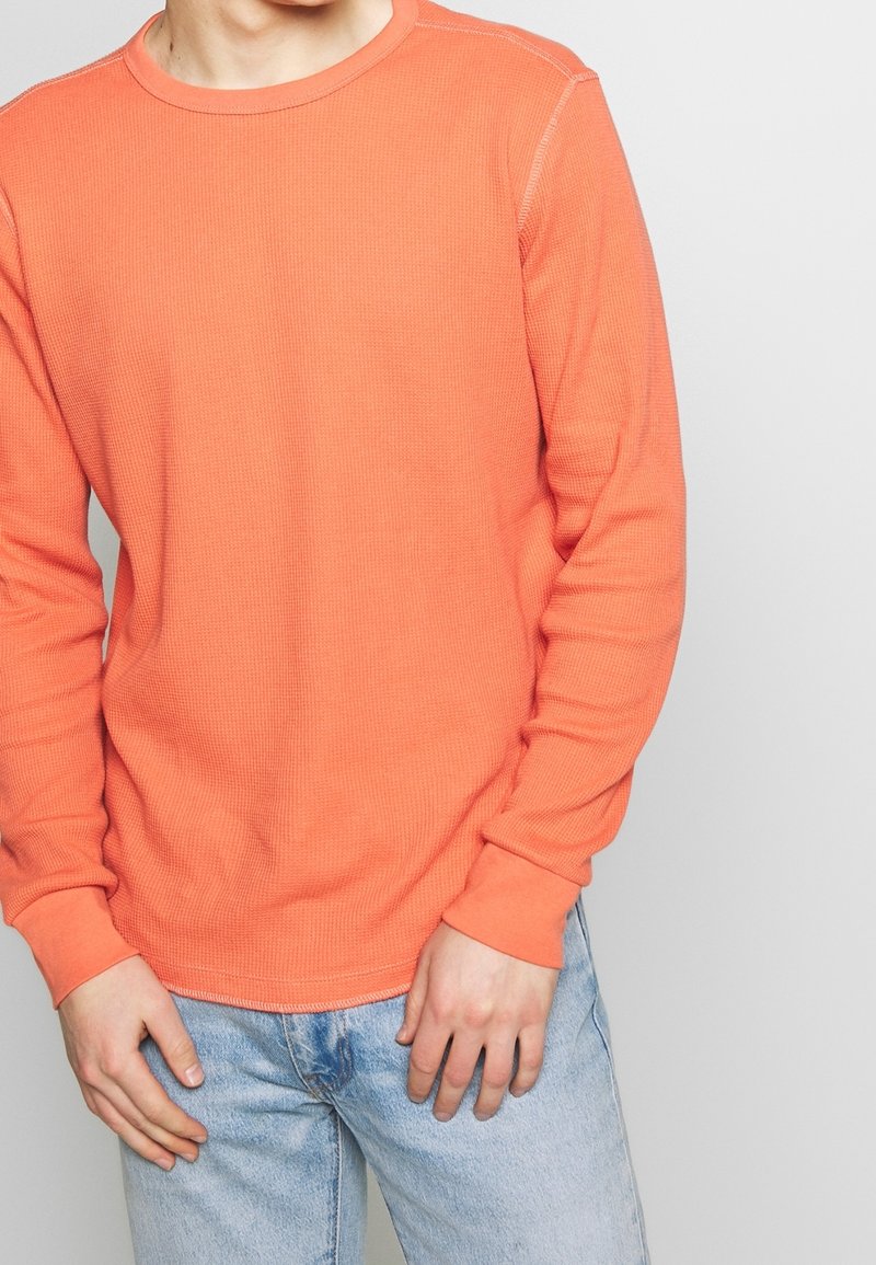 Chemise à manches longues en tissu gaufré de couleur orange vif, avec un col rond et des poignets ajustés, accompagnée de jeans en denim bleu clair.