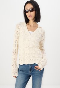 Helle, cremefarbene Strickjacke mit gewelltem Saum, die offene florale Muster und lange Ärmel aufweist, kombiniert mit blauen Jeans.