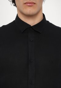 Μαύρο πουκάμισο με κουμπιά και δομημένο γιακά, φτιαγμένο από υφασμάτινη υφή, διακοσμημένο με μια διακριτική λογότυπο και τυπικό κλείσιμο με κουμπιά.