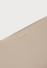 Prodotto in pelle beige con texture e logo "BOSS" dorato. Presenta linee pulite e un design minimalista, che mette in risalto il suo aspetto sofisticato.