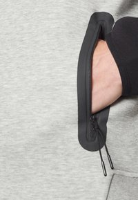 Grå sweatshirtstof med en sort lynlåslomme. Lynlåsen har sorte trækstropper, og lommen er integreret i hætten.