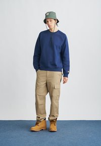 Μπλε ναυτικό φούτερ, μπεζ παντελόνι cargo με τσέπες, πράσινο καπέλο τύπου bucket και καφέ μπότες, στέκεται σε μπλε χαλί με απλό λευκό φόντο.