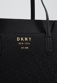 Sac cabas noir DKNY New York avec poignées en cuir texturé et lettres dorées indiquant "DKNY New York EST. 1989" sur tissu à motifs.