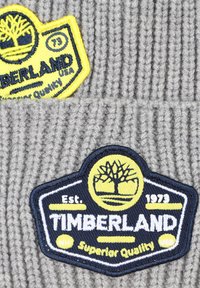 Tissu en tricot gris avec deux écussons brodés de la marque Timberland en jaune, bleu marine et blanc, comportant un logo d'arbre et des détails textuels.