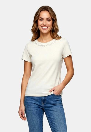 Donna sorridente con capelli ondulati, indossa una maglietta bianca a maniche corte con testo sul colletto e jeans blu, mano in tasca, sfondo grigio chiaro.