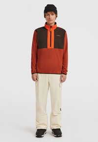 Fleece-Pullover in Orange und Braun mit Frontreißverschluss, hohem Kragen und Logo. Kombiniert mit hellbeigen Schneehosen und schwarzen Stiefeln.