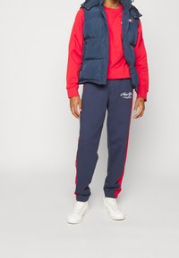 Granatowa puchowa kamizelka, czerwona koszula z długim rękawem, granatowe dresy z czerwonymi bocznymi paskami oraz białe trampki. Na koszuli i spodniach logo marki.