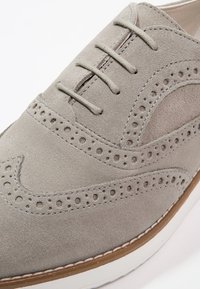 Gros plan d’une chaussure à bout golf en daim gris clair avec perforations brogue, lacets assortis et semelle blanche avec bordure marron.