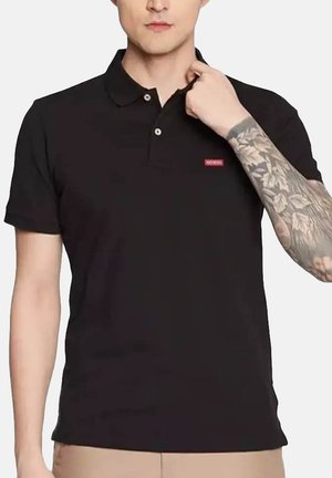 Homme portant un polo noir à manches courtes avec un petit logo rouge, ajustant le col et montrant une manche de tatouage florale sur le bras droit.